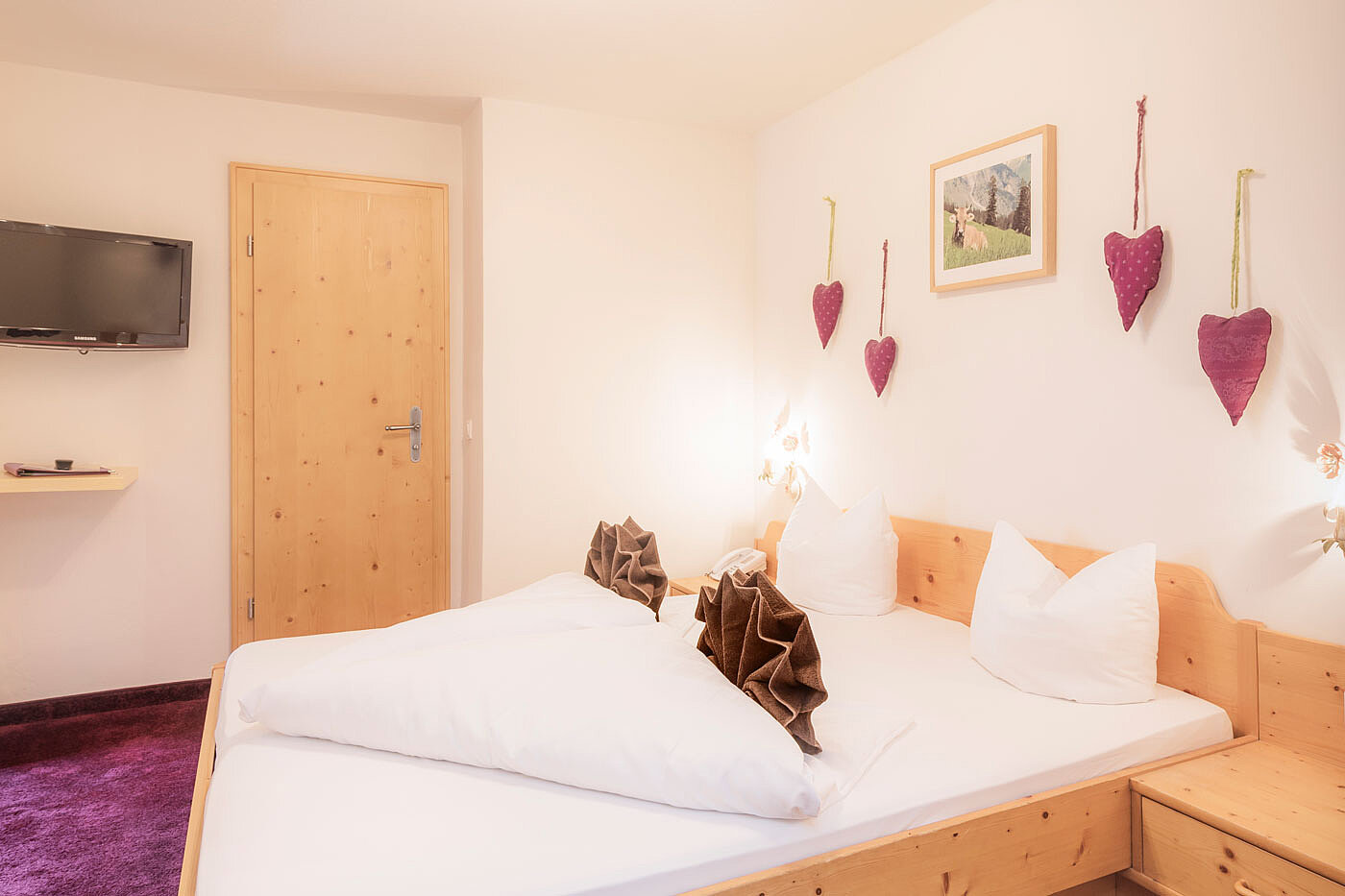Gemütliches Hotelzimmer mit Holzbett, lila Teppich, Wandfernseher und herzförmiger Wanddekoration.