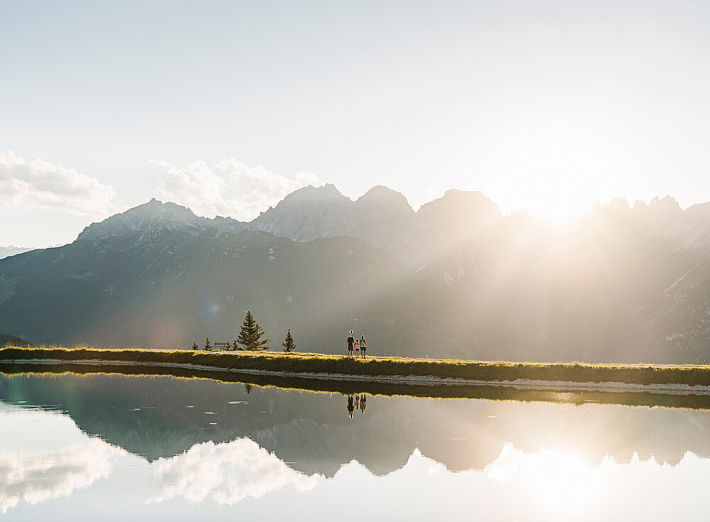 Die Sonne geht über den Bergen auf und spiegelt sich in einem ruhigen See, an dessen grasbewachsenem Ufer Menschen entlanggehen.