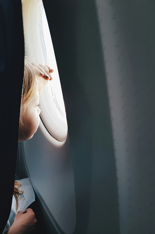 Ein Kind schaut aus dem Flugzeugfenster, hält ein Stück Papier in der Hand und das Sonnenlicht beleuchtet sein Haar.