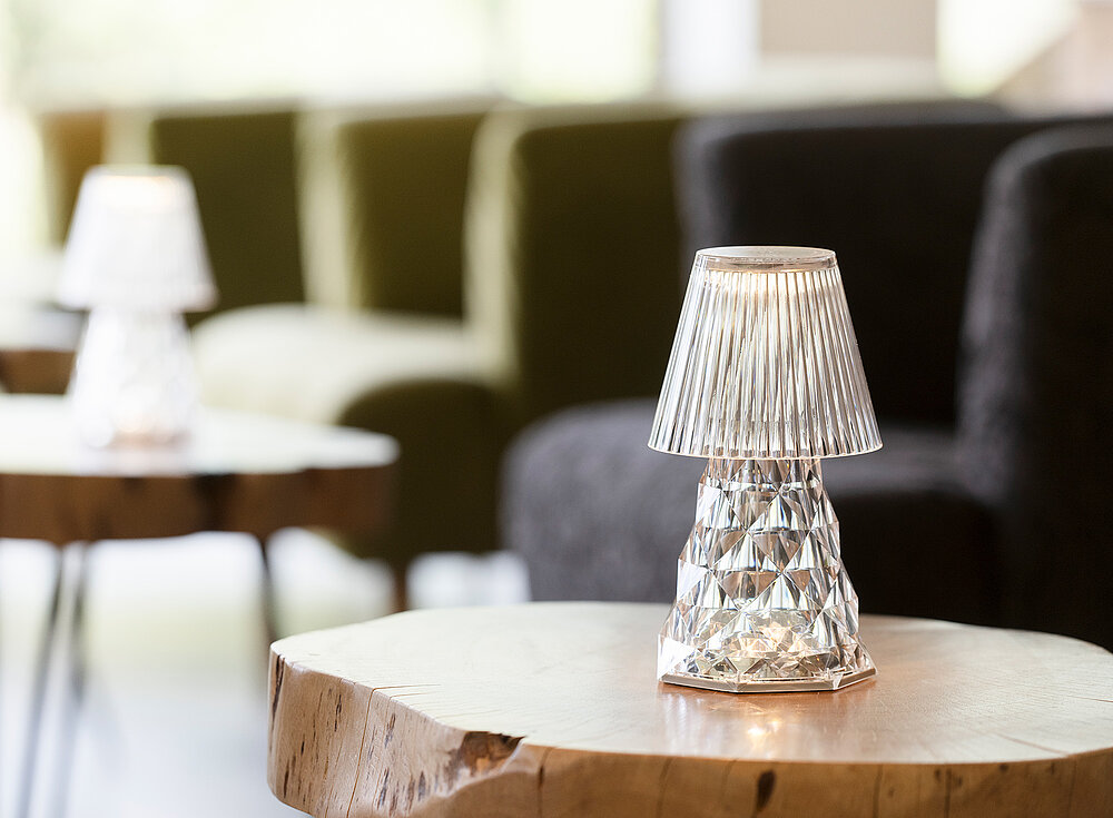 Eine kleine, moderne Tischlampe steht auf einem Holztisch mit verschwommenen Stühlen im Hintergrund.