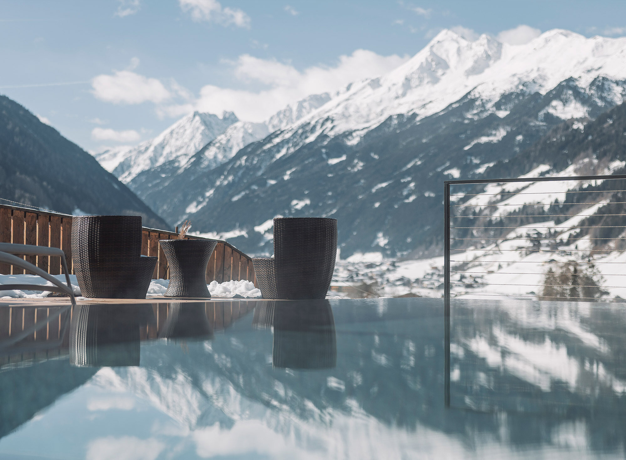 Die Sitzgelegenheiten im Freien mit schwarzen Korbstühlen auf der Terrasse eines Hotels in Neustift bieten einen Blick auf die schneebedeckten Berge des Stubaitals, deren Gipfel und Himmel sich im Infinity-Pool darunter spiegeln. Die ruhige Szene wird von einem Holzgeländer und Metallseilen eingerahmt.