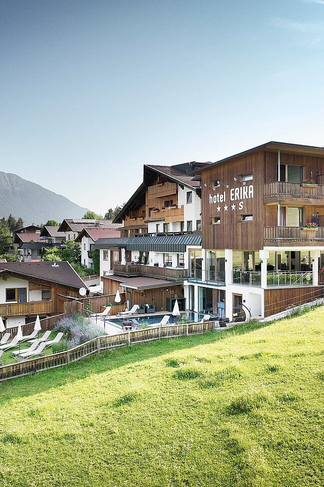 Das Hotel Erika mit viel Holz in Neustift liegt auf einem grünen Hügel und bietet unter klarem Himmel einen Blick auf die Berge des Stubaitals.