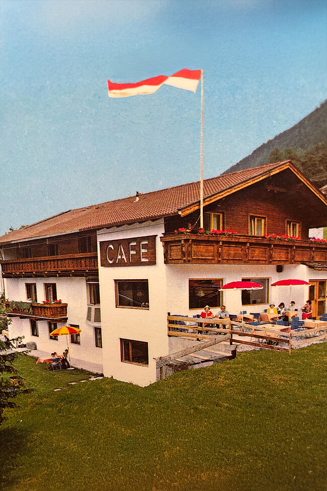 Café im alpenländischen Stil mit Balkon, Sitzgelegenheiten im Freien und einer österreichischen Flagge, umgeben von einer grünen Wiese und Bergen.