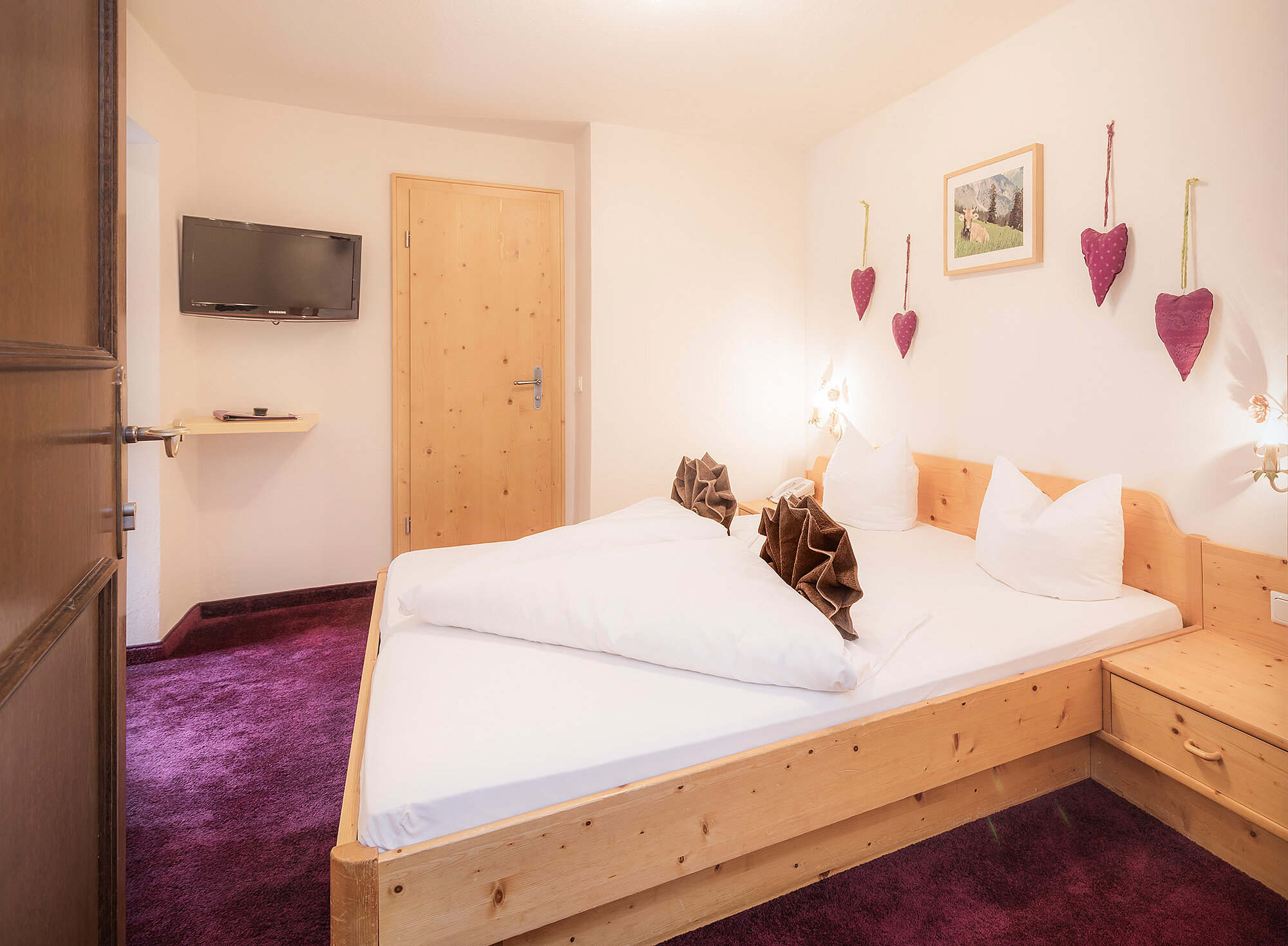 Gemütliches Hotelzimmer mit Holzbett, lila Teppich, Wandfernseher und herzförmiger Wanddekoration.
