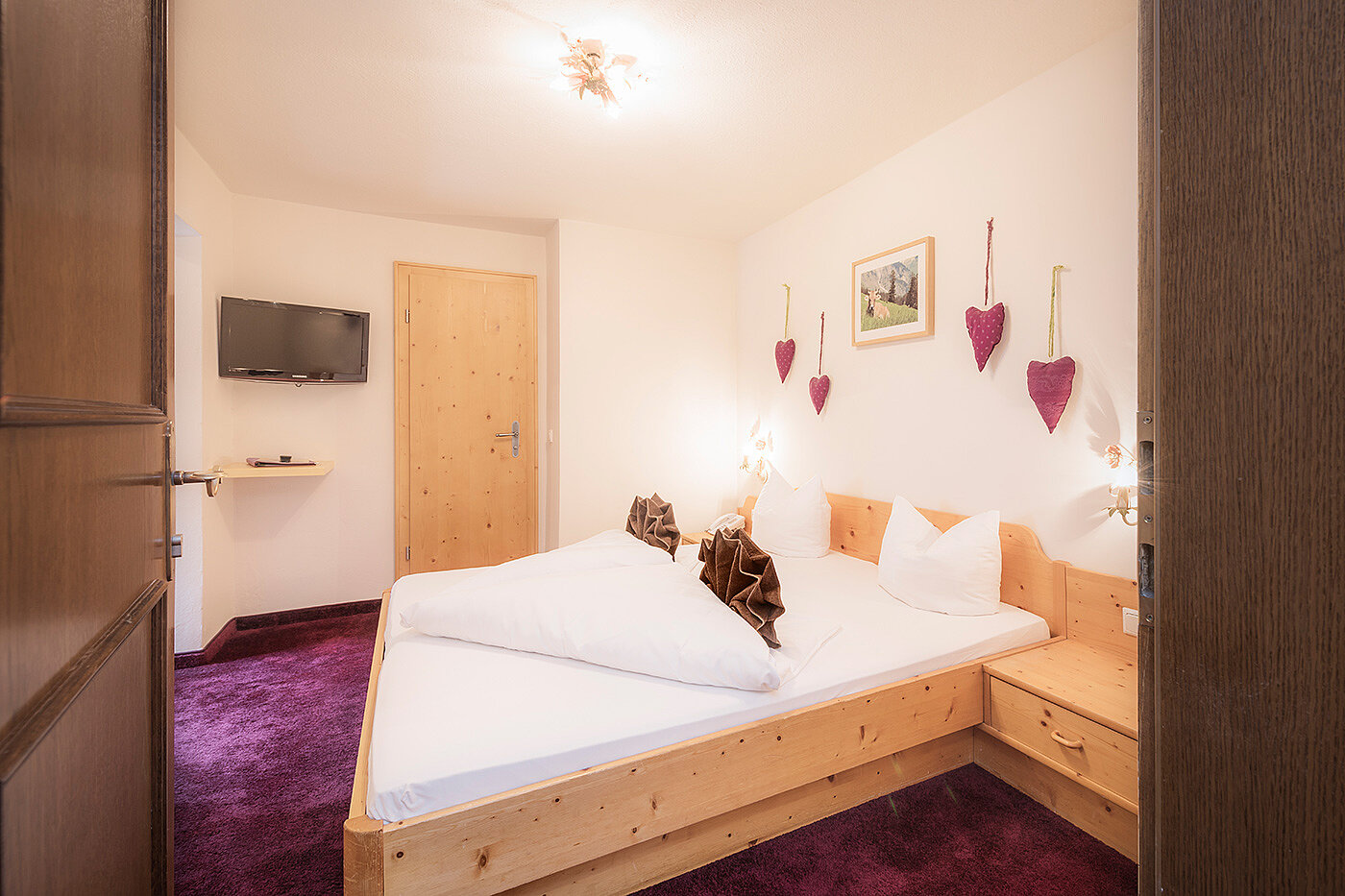 Gemütliches Hotelzimmer mit Doppelbett, Holzmöbeln, Wandfernseher und lila Teppich. Herzdekorationen an der Wand.