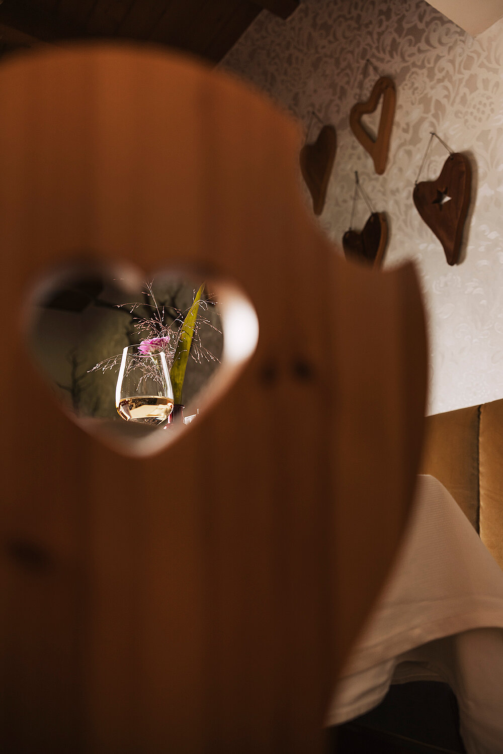 Durch einen herzförmigen Ausschnitt in einem Holzstuhl ist ein Blumenstrauß zu sehen, während weitere Herzformen die Wand im Hintergrund zieren.