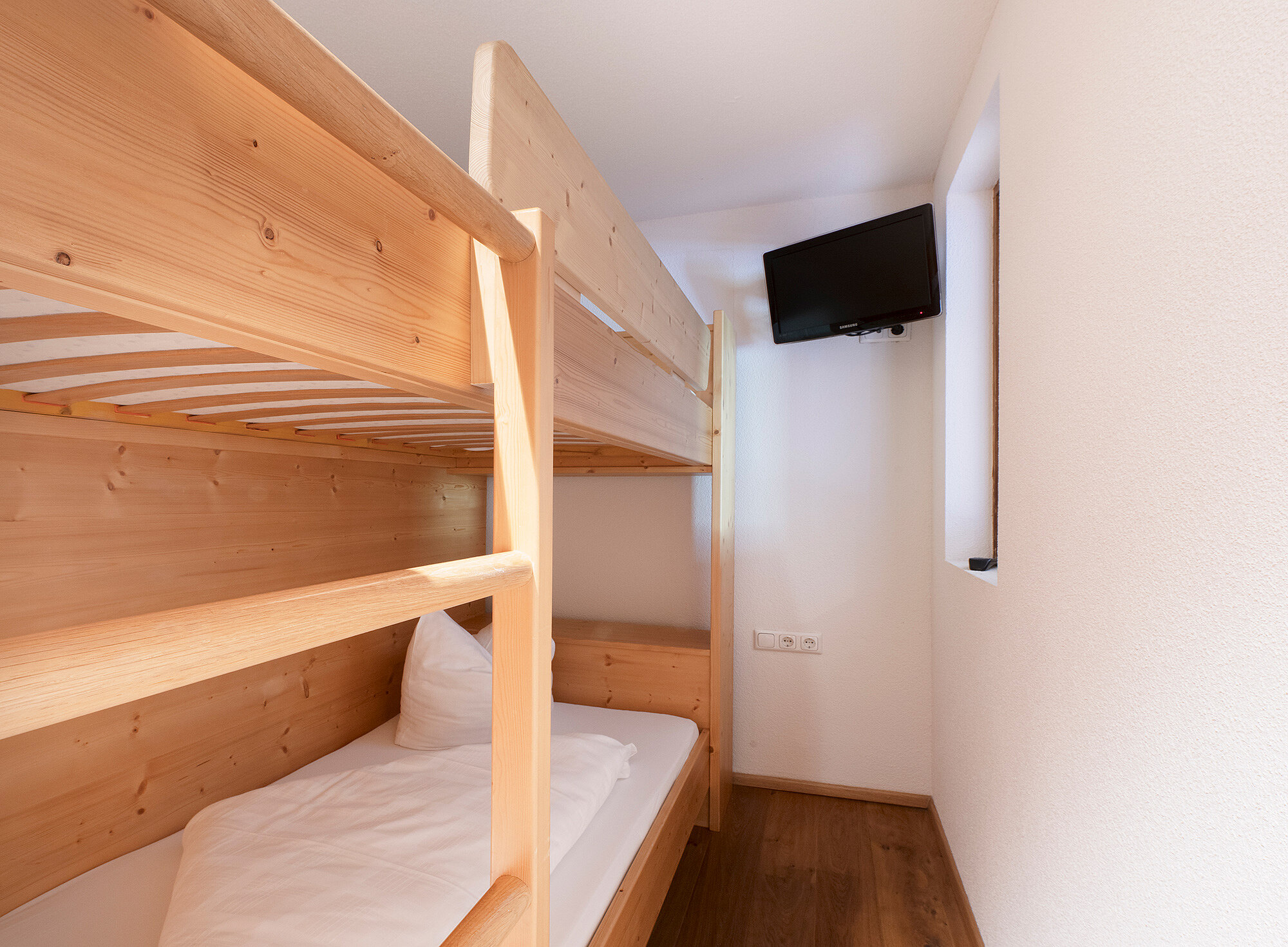 Schmales Zimmer mit Etagenbetten aus Holz, weißen Wänden, einem Fenster und einem an der Wand montierten Fernseher.