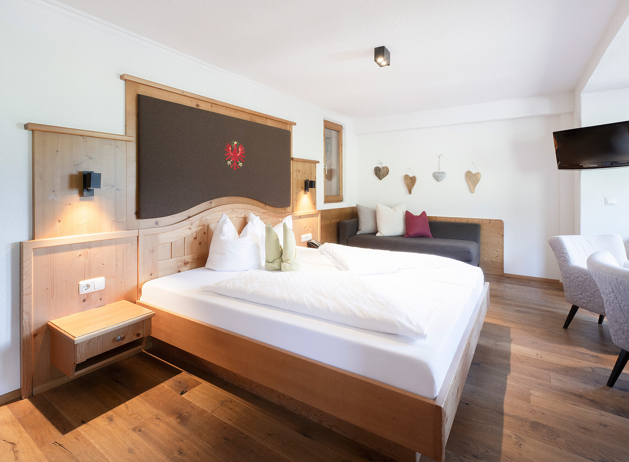 Helles Hotelzimmer mit großem Bett, Holzakzenten, Sofa, Wanddekor und Fernseher auf der rechten Seite.