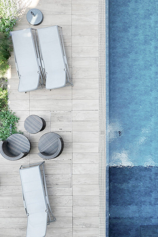 Luftaufnahme des Poolbereichs mit zwei Liegestühlen, drei runden Tischen und Pflanzen neben einem blauen Swimmingpool.
