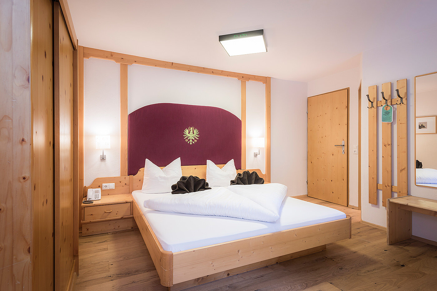 Ein gemütliches Hotelzimmer mit einem Doppelbett, Holzmöbeln, Wandlampen und einem Spiegel neben der Tür.