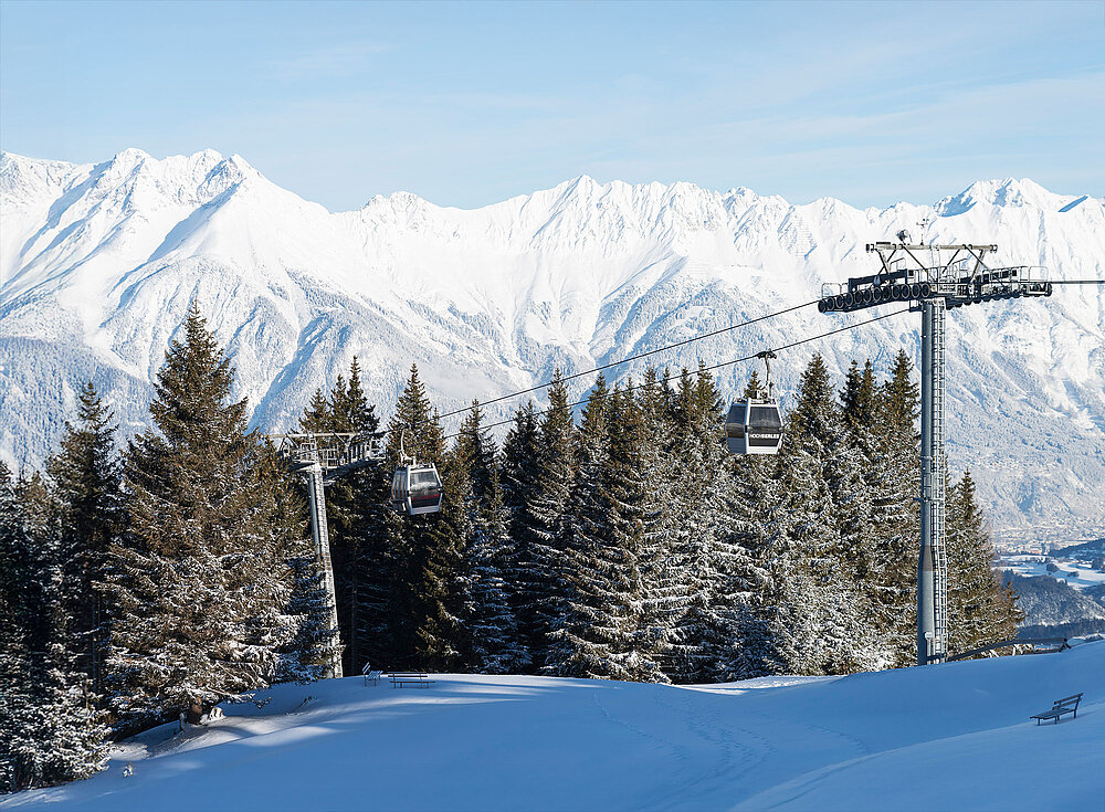 Verschneite Berglandschaft mit Skilift, Kiefern im Vordergrund und strahlend blauem Himmel im Hintergrund.