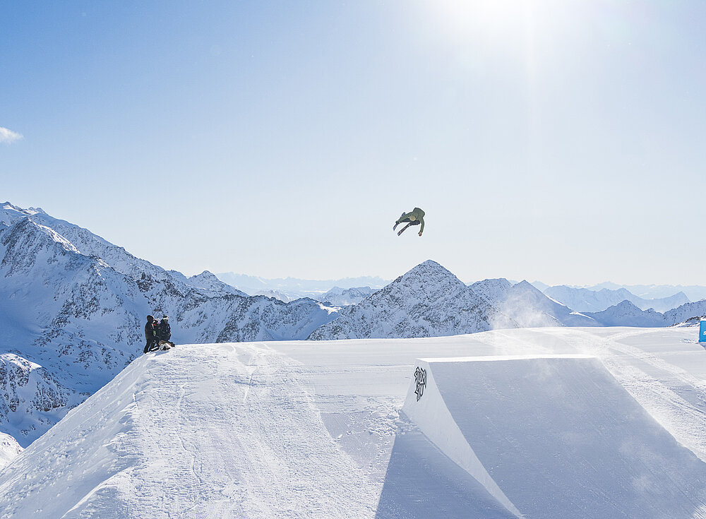 Ein Snowboarder macht einen Sprung auf einem schneebedeckten Berghang unter einem hellen, klaren Himmel mit Blick auf die Gipfel in der Ferne.