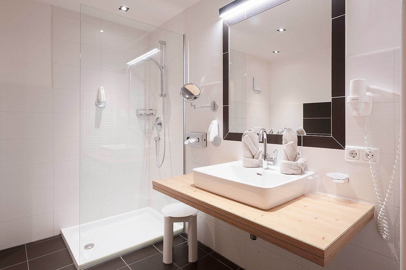 Modernes Badezimmer mit schwarz-weißen Fliesen, schwebender Toilette, ebenerdiger Dusche, Waschbecken, großem Spiegel und heller Beleuchtung.