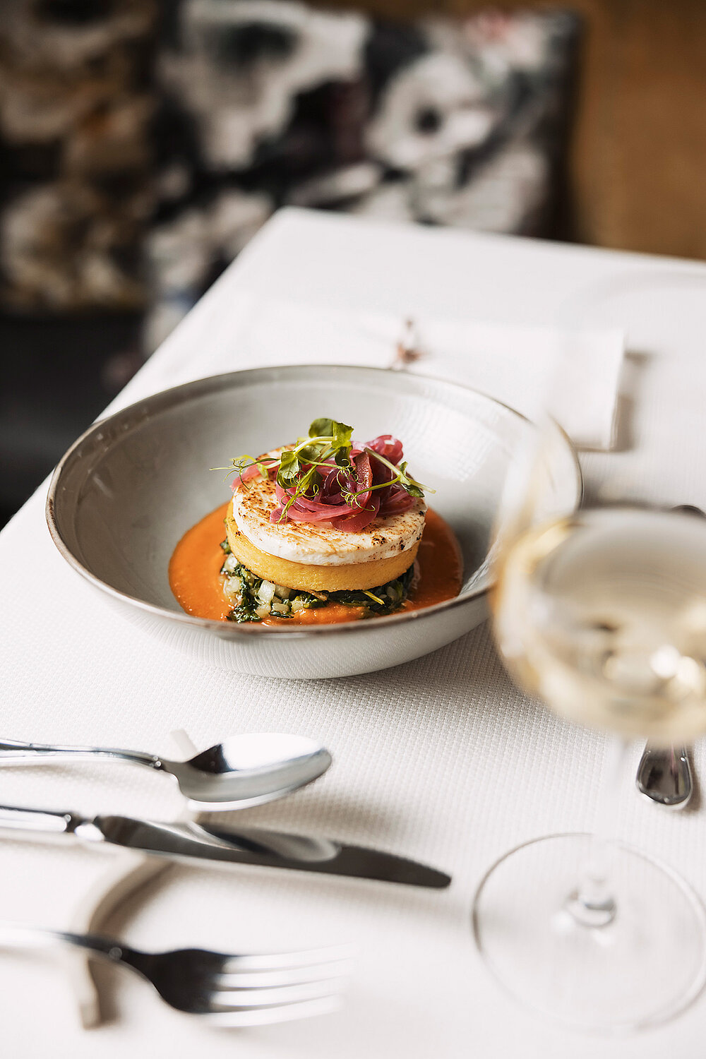 Ein Gourmetgericht mit einer runden, vielschichtigen Hauptspeise, garniert mit Microgreens, steht in einer grauen Schüssel mit orangefarbener Soße auf einer weißen Tischdecke neben Wein, Gabel, Messer und Serviette.