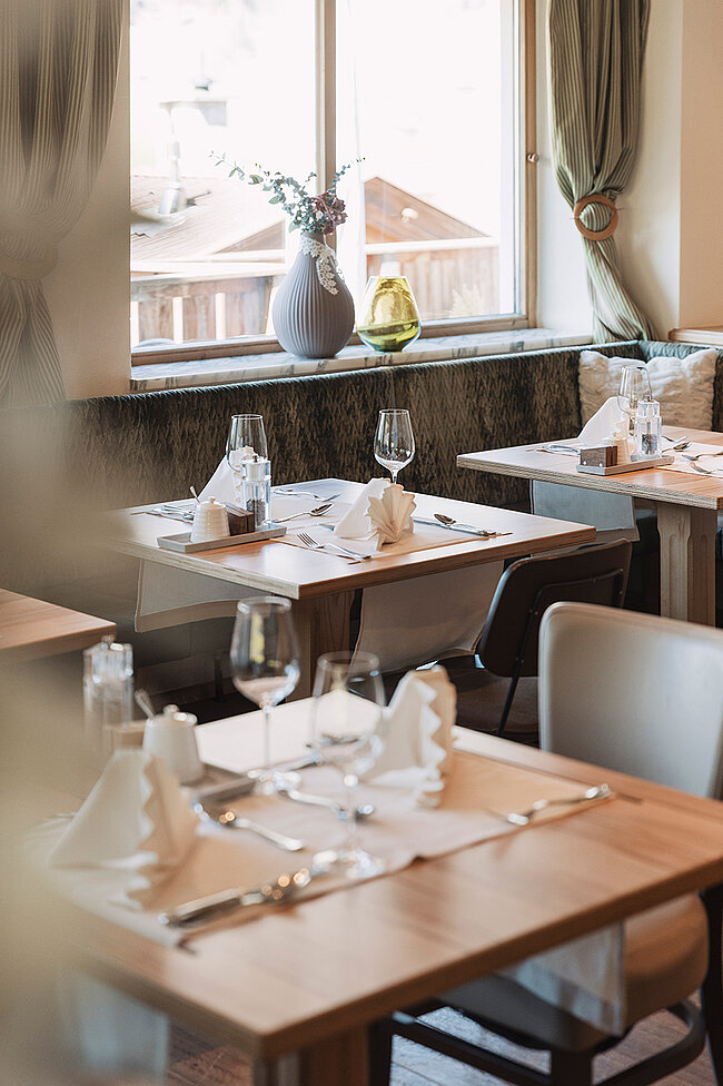 Gemütlicher Restauranttisch mit Gläsern, Tellern und Servietten in der Nähe eines Fensters mit Tageslicht und dekorativer Vase.