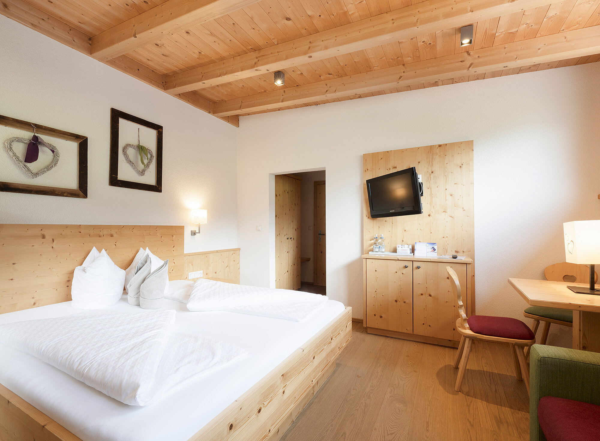 Helles Hotelzimmer mit Holzmöbeln, Doppelbett, an der Wand montiertem Fernseher, Schreibtisch, Stühlen und gerahmter Kunst an der Wand.