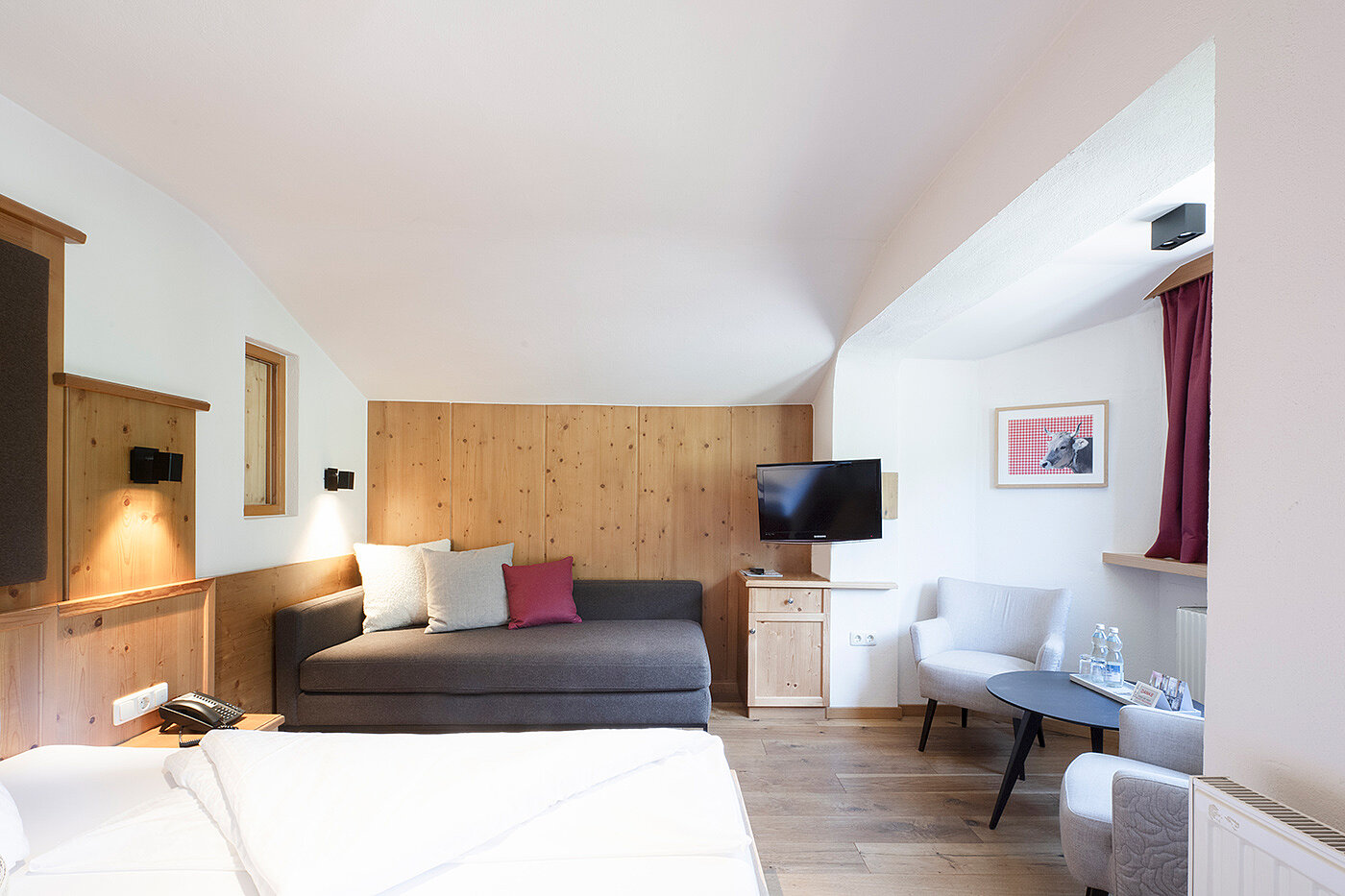 Helles, modernes Hotelzimmer mit Bett, Sofa, Fernseher, zwei Stühlen, kleinem Tisch und Holzakzenten.