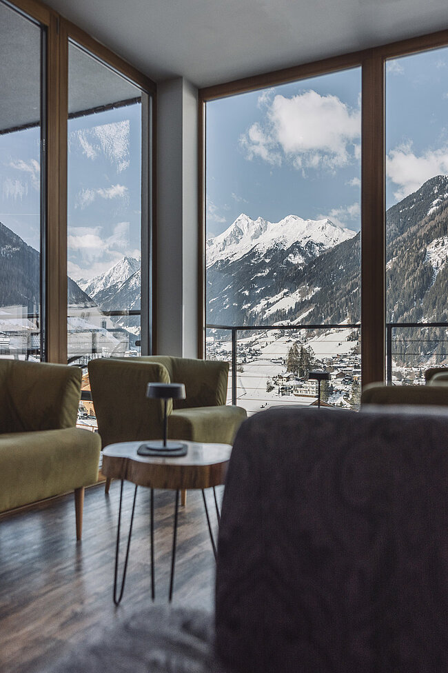 Ein moderner Aufenthaltsraum in einem Hotel in Neustift mit olivgrünen Stühlen und einem kleinen Tisch. Die großen Fenster bieten einen malerischen Blick auf die schneebedeckten Berge und das Stubaital unter einem teilweise bewölkten Himmel.