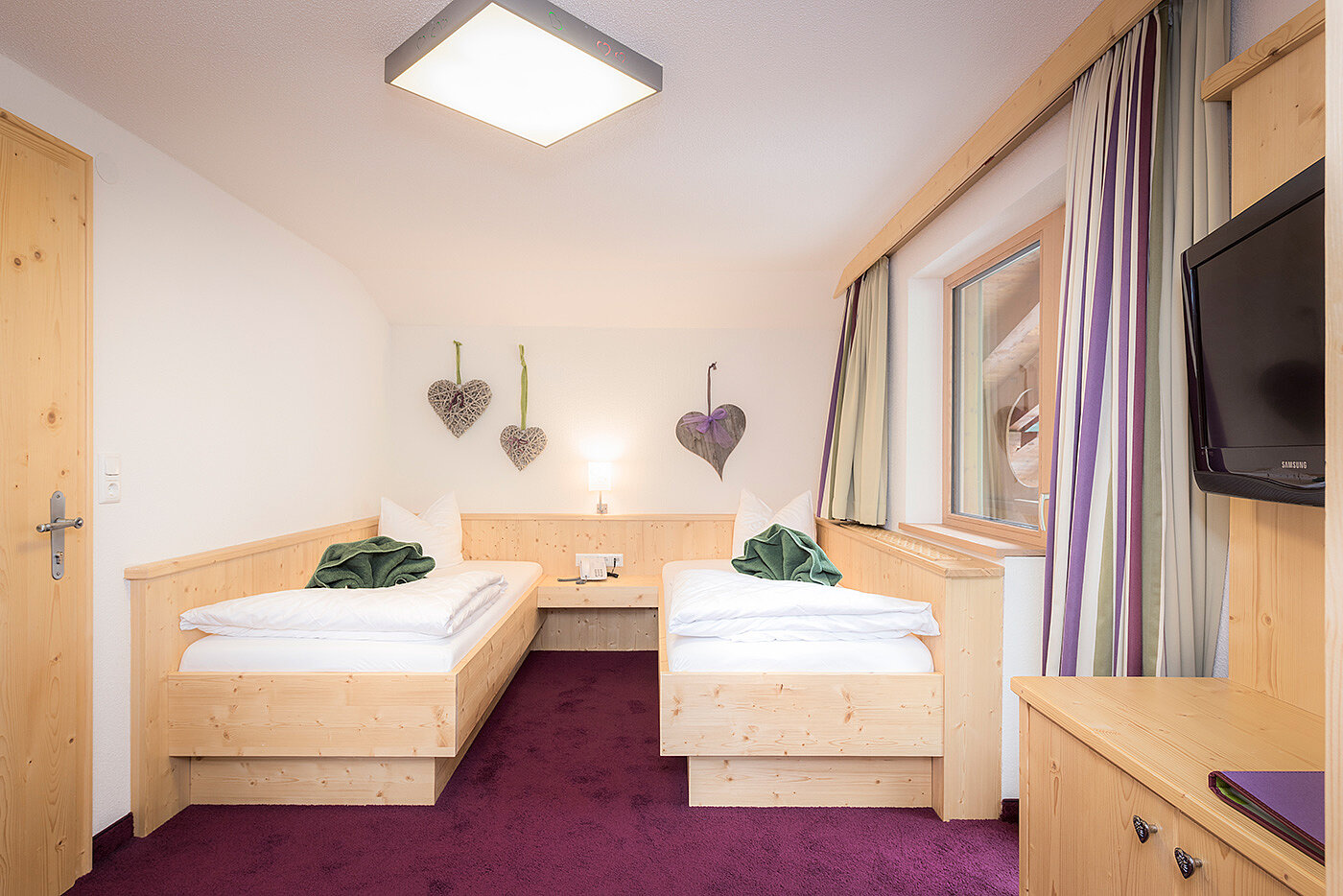 Gemütliches Hotelzimmer mit zwei Einzelbetten, Holzmöbeln, lila Teppich und herzförmiger Wanddekoration.