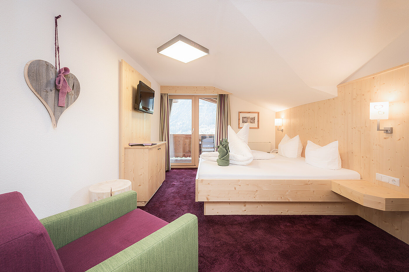 Modernes Hotelzimmer mit Doppelbett, grünem Sofa, herzförmiger Wanddekoration und sanfter Beleuchtung.