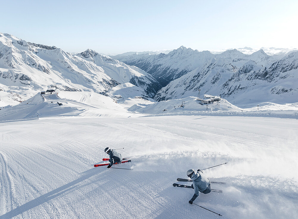 Zwei Skifahrer ziehen unter klarem Himmel ihre Schwünge auf einem breiten, verschneiten Hang, umgeben von schneebedeckten Bergen.