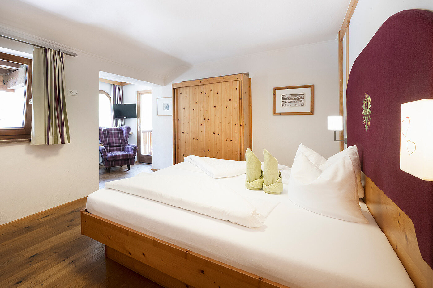 Helles Hotelzimmer mit Holzbett, Kleiderschrank, weißer Bettwäsche und einem Fenster mit Tageslicht.