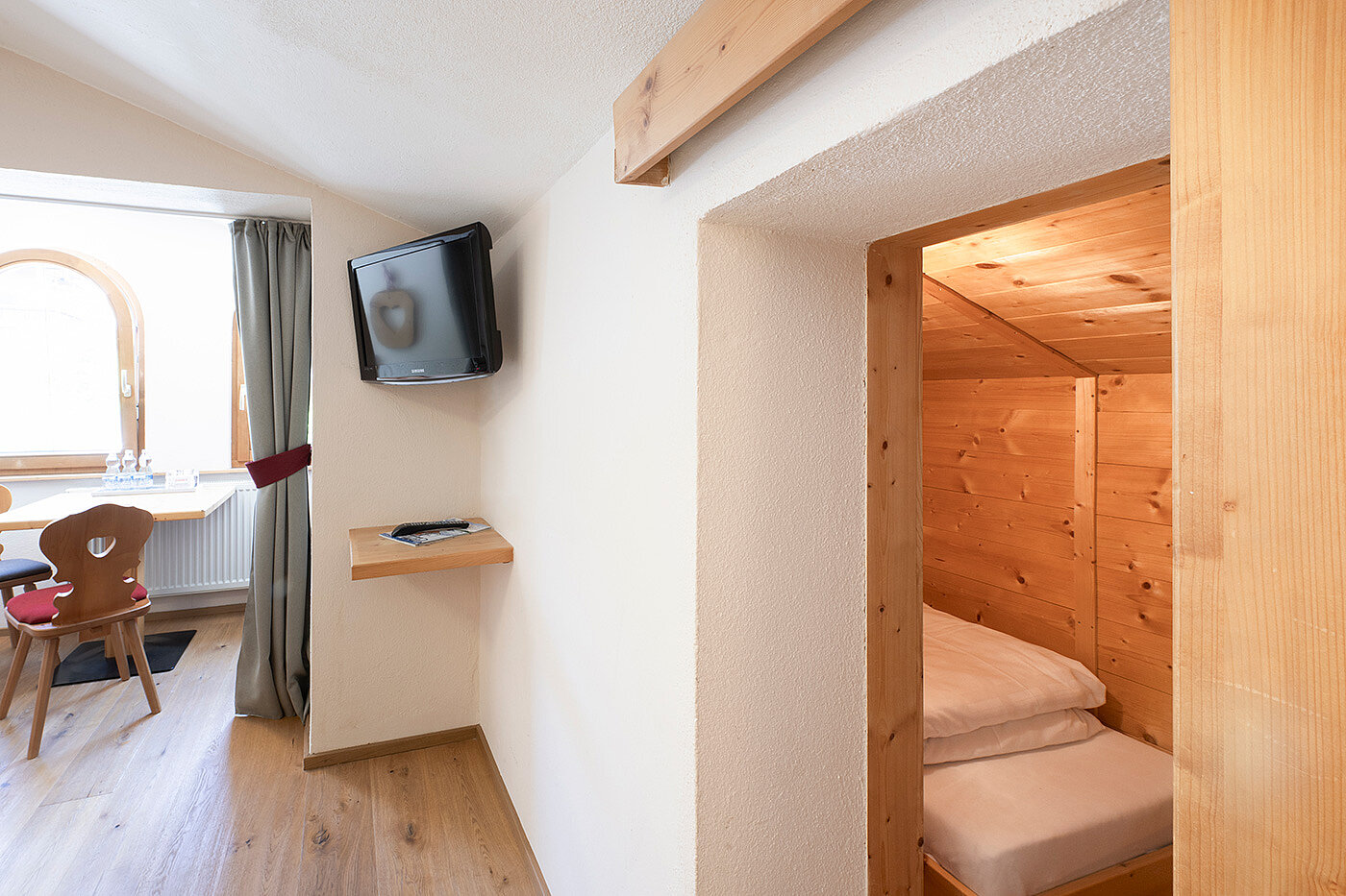 Kleines, gemütliches Zimmer mit an der Wand montiertem Fernseher, Schreibtisch und holzgetäfelter Schlafecke mit ordentlich gemachtem Bett.