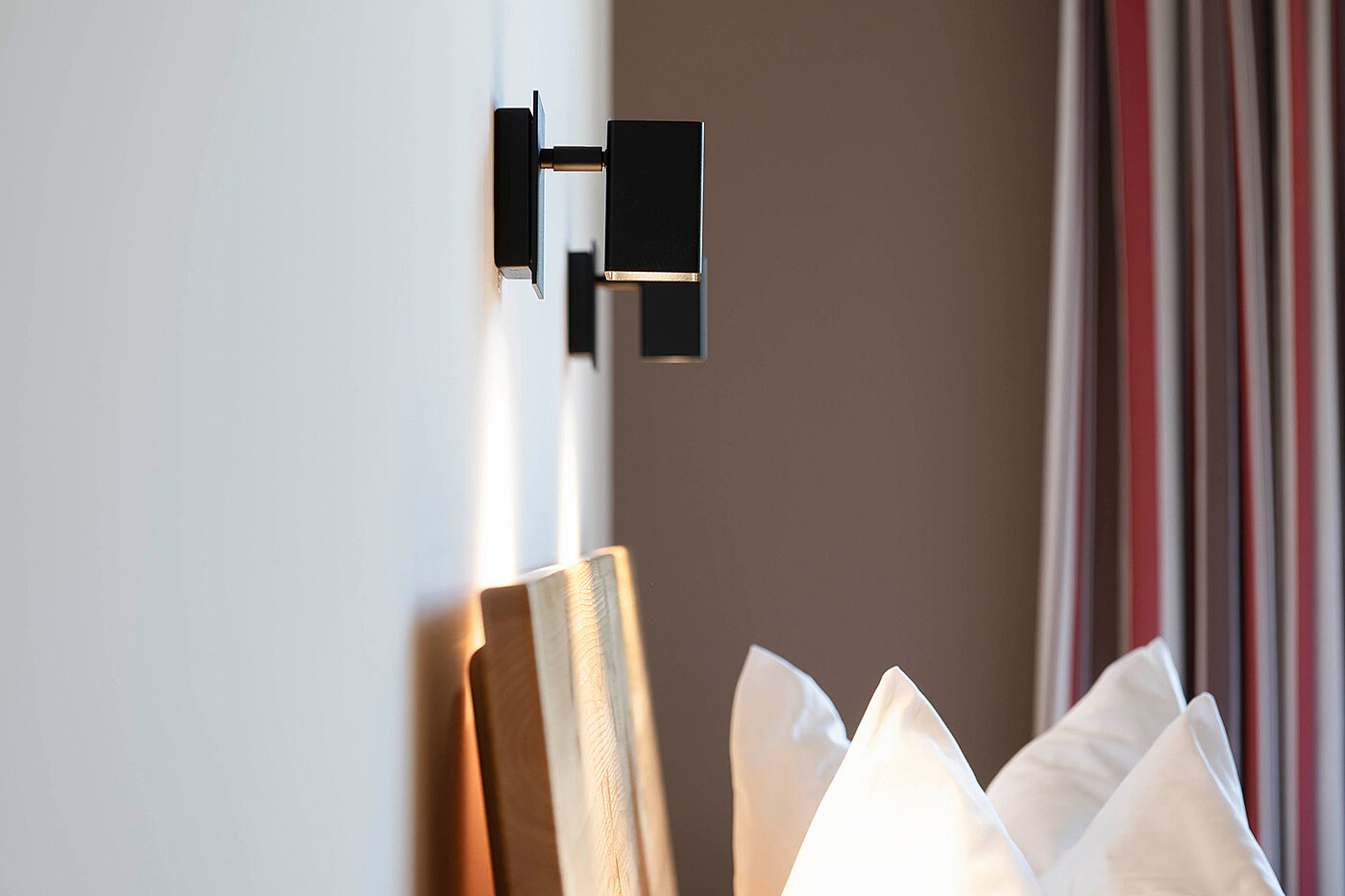 Moderne Wandlampe über einem hölzernen Kopfteil und weißen Kissen, mit gestreiften Vorhängen im Hintergrund.