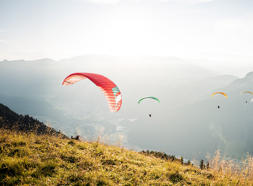 Vier Gleitschirmflieger schweben über einem grasbewachsenen Hügel mit Bergen und einem dunstigen Himmel im Hintergrund.