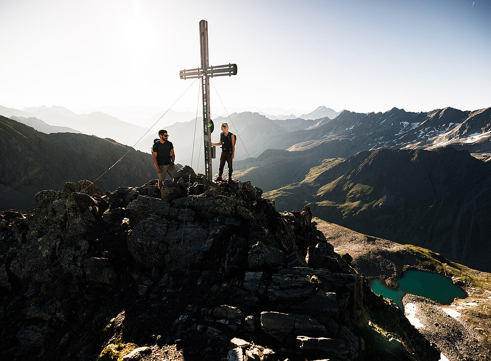 Zwei Wanderer stehen neben einem großen Kreuz auf einem felsigen Berggipfel. Unter ihnen sind im Sonnenlicht Täler und Seen sichtbar.