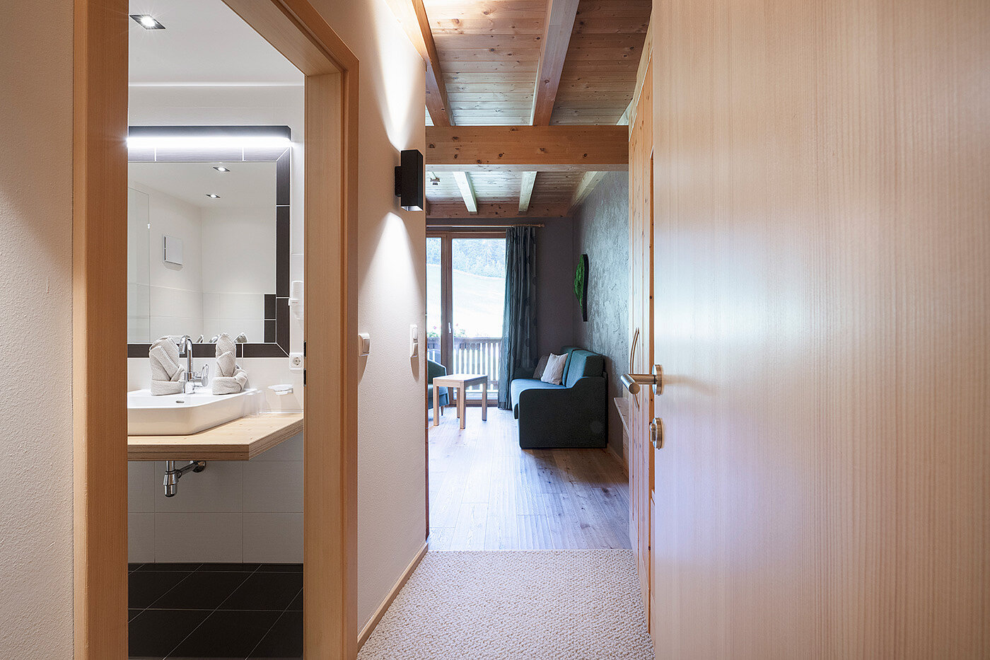 Blick von einem Flur in ein modernes Badezimmer auf der linken Seite und einen Wohnbereich mit einem Sofa und einem großen Fenster weiter vorne.