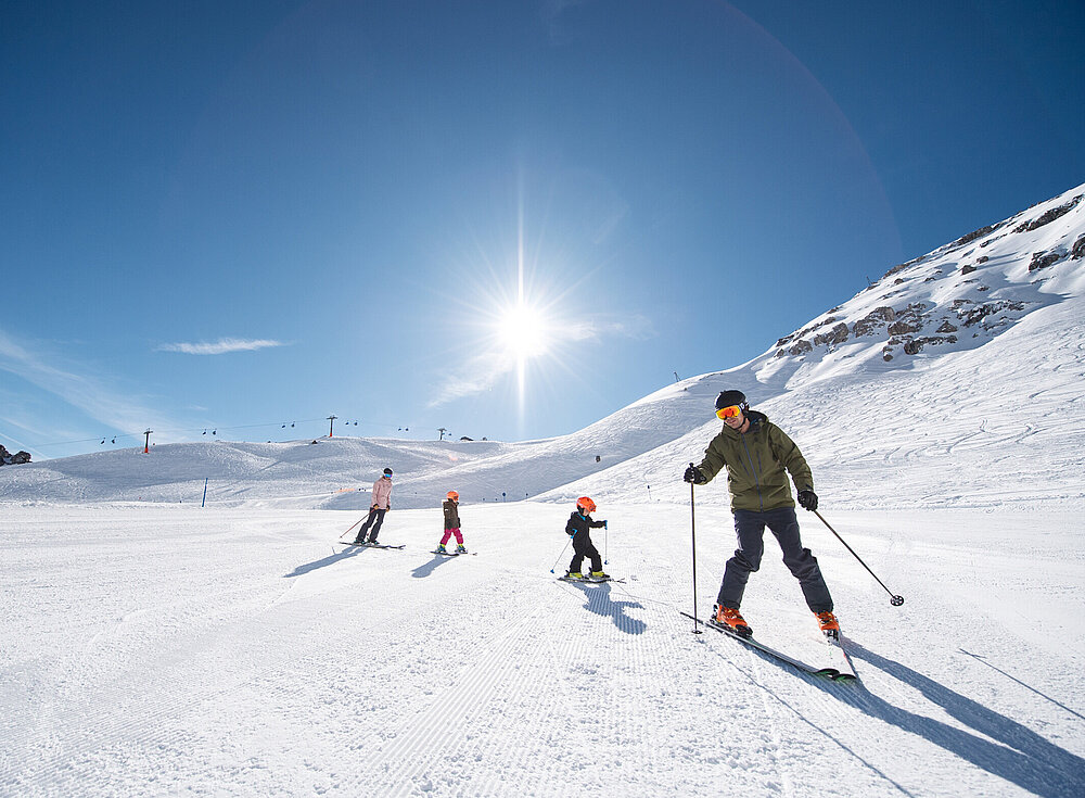 Vier Personen fahren unter strahlender Sonne und klarem blauen Himmel Ski auf einem schneebedeckten Hang, im Hintergrund sind Berge zu sehen.