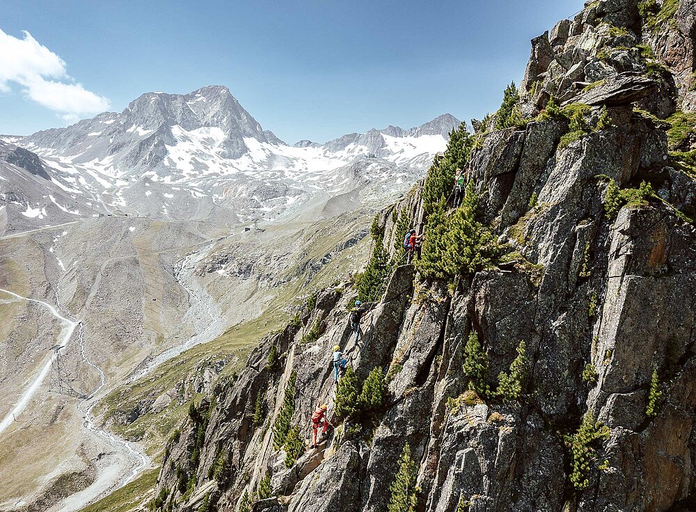 Ein Bergsteiger erklimmt unter klarem Himmel einen felsigen, baumbestandenen Berghang mit schneebedeckten Gipfeln im Hintergrund.