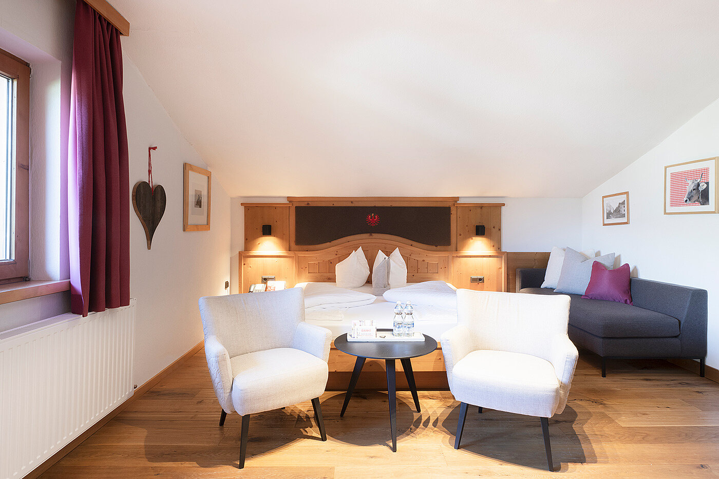 Gemütliches Hotelzimmer mit einem Doppelbett, zwei weißen Sesseln, einem kleinen runden Tisch und einem dunklen Sofa auf Holzboden.
