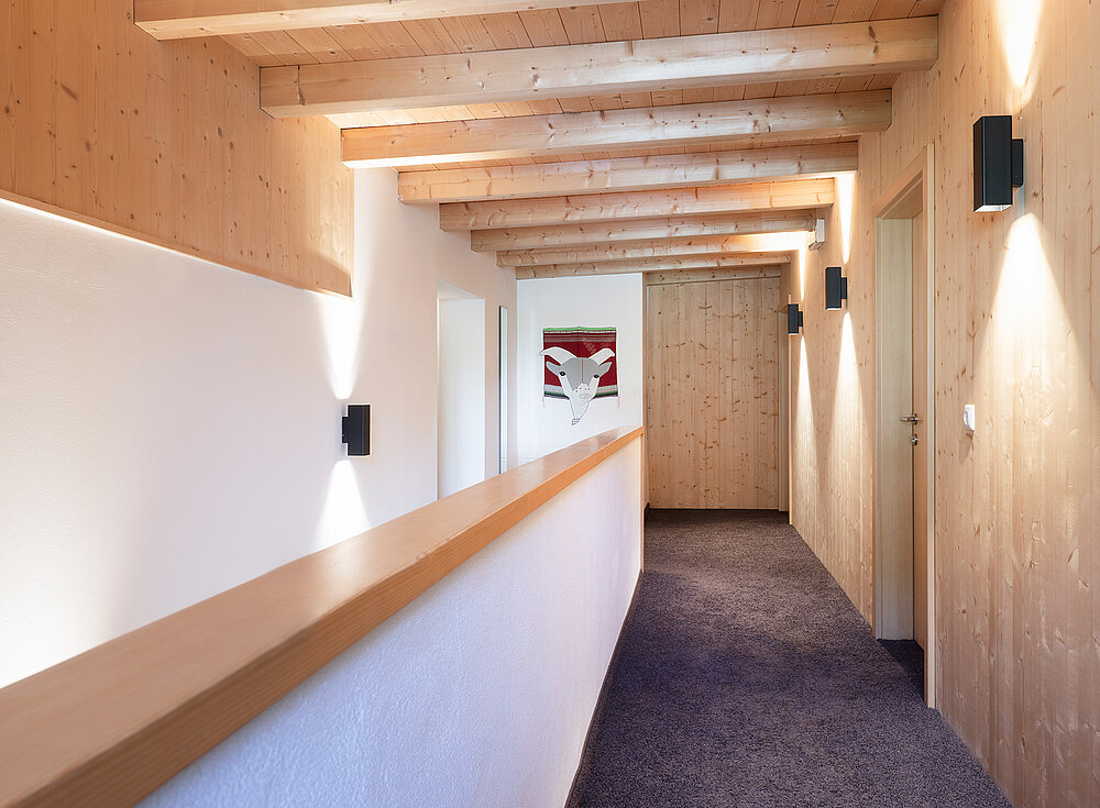 Ein moderner Flur mit Holzbalken, weißen Wänden und einer geschlossenen Holztür am Ende; an der linken Wand hängt ein Gemälde.
