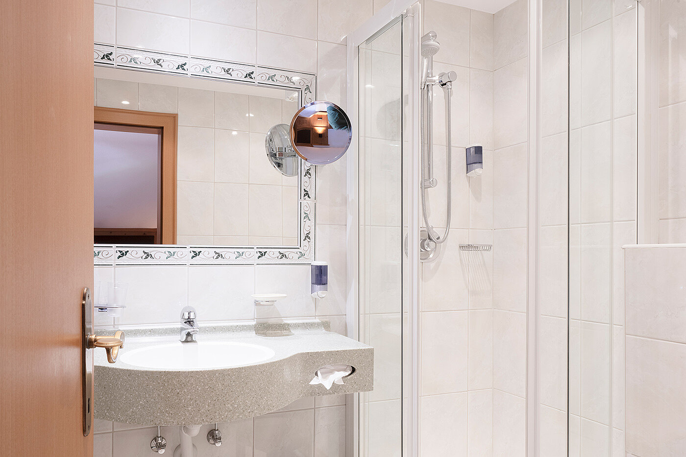 Weiß gefliestes Badezimmer mit Waschbecken, Wandspiegel, Duschkabine und einem Holzrahmenfenster an der linken Wand.