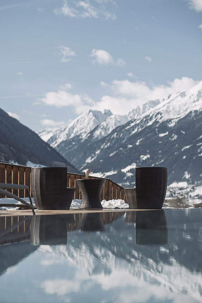 Drei Korbstühle und ein runder Tisch stehen auf der Terrasse eines Hotels in Neustift, mit Blick auf die schneebedeckten Berge des Bergblicks, deren Spiegelung unter dem teilweise bewölkten Himmel des Stubaitals auf einer glänzenden Oberfläche schimmert.