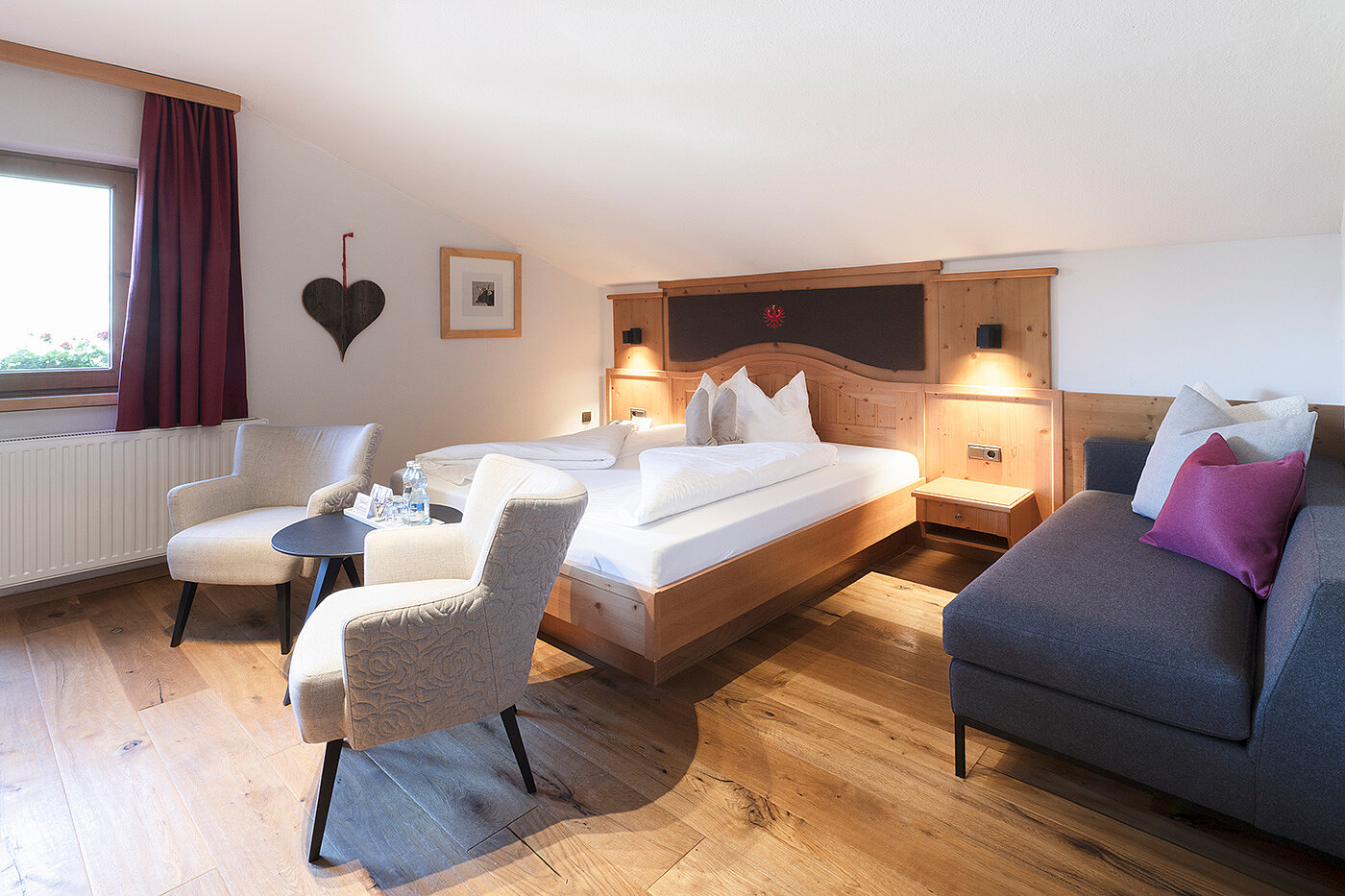 Gemütliches Hotelzimmer mit einem Doppelbett, zwei Sesseln, einem kleinen runden Tisch, einem Sofa und Holzboden.