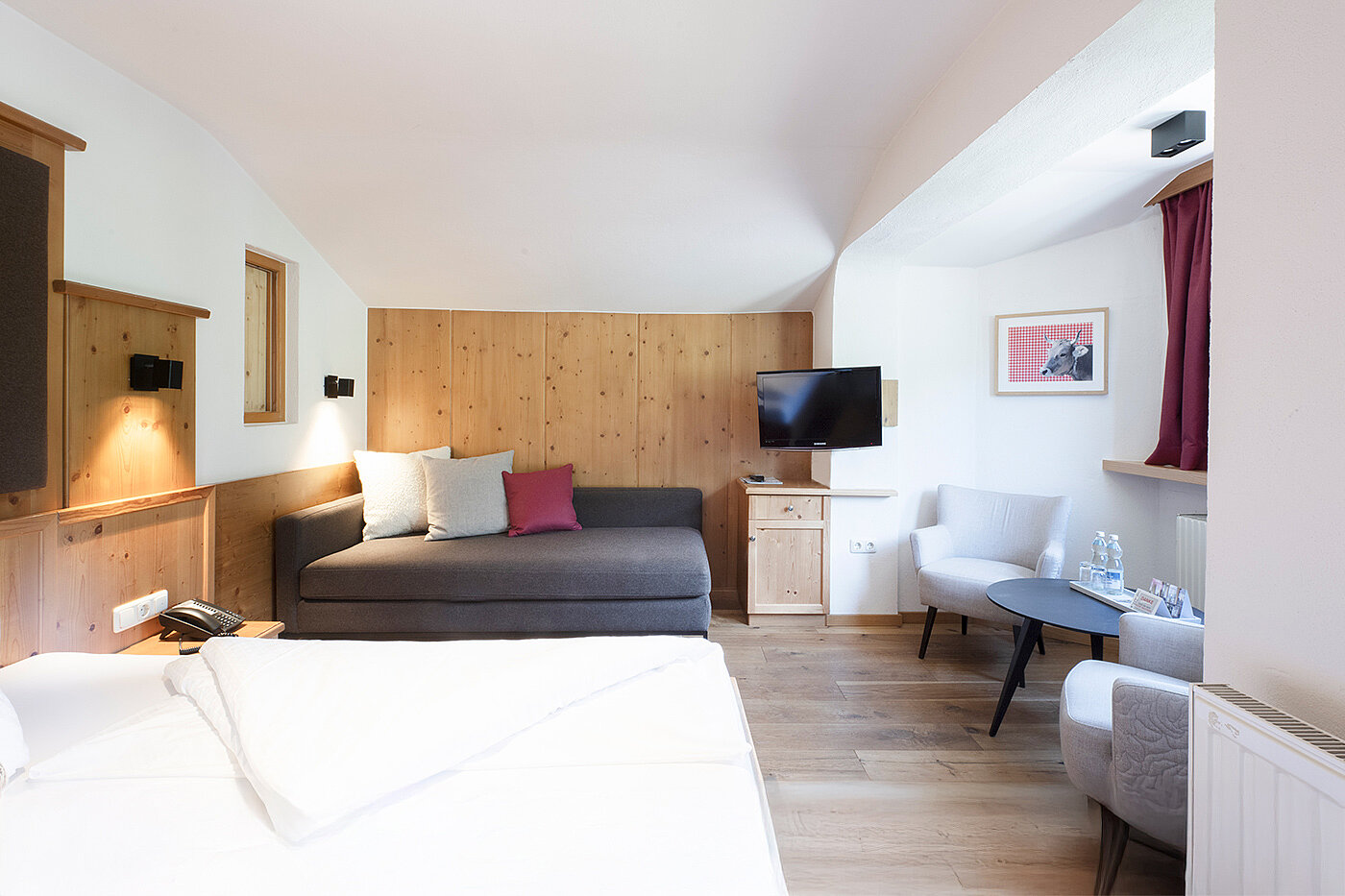 Modernes Hotelzimmer mit Bett, Sofa, Fernseher, Stühlen, Tisch, Holzakzenten und natürlichem Licht durch die Fenster.
