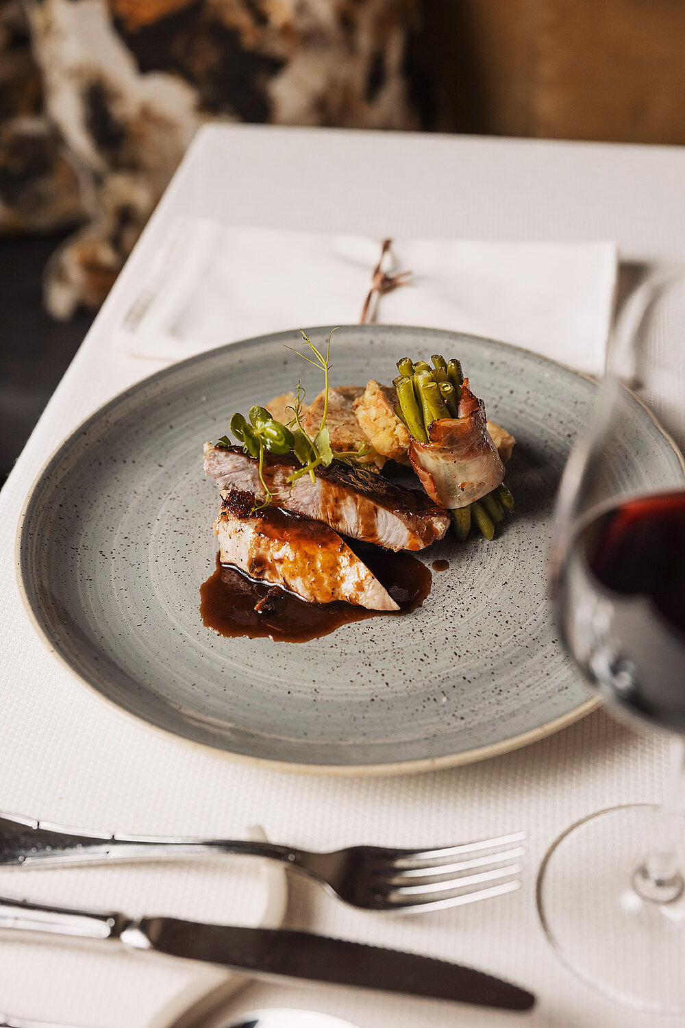 Ein ansprechend angerichtetes Gourmetgericht mit Fleisch, Soße und Gemüse auf einem grauen Teller steht auf einer weißen Tischdecke neben einem Glas Rotwein, Besteck und einer gefalteten Serviette.