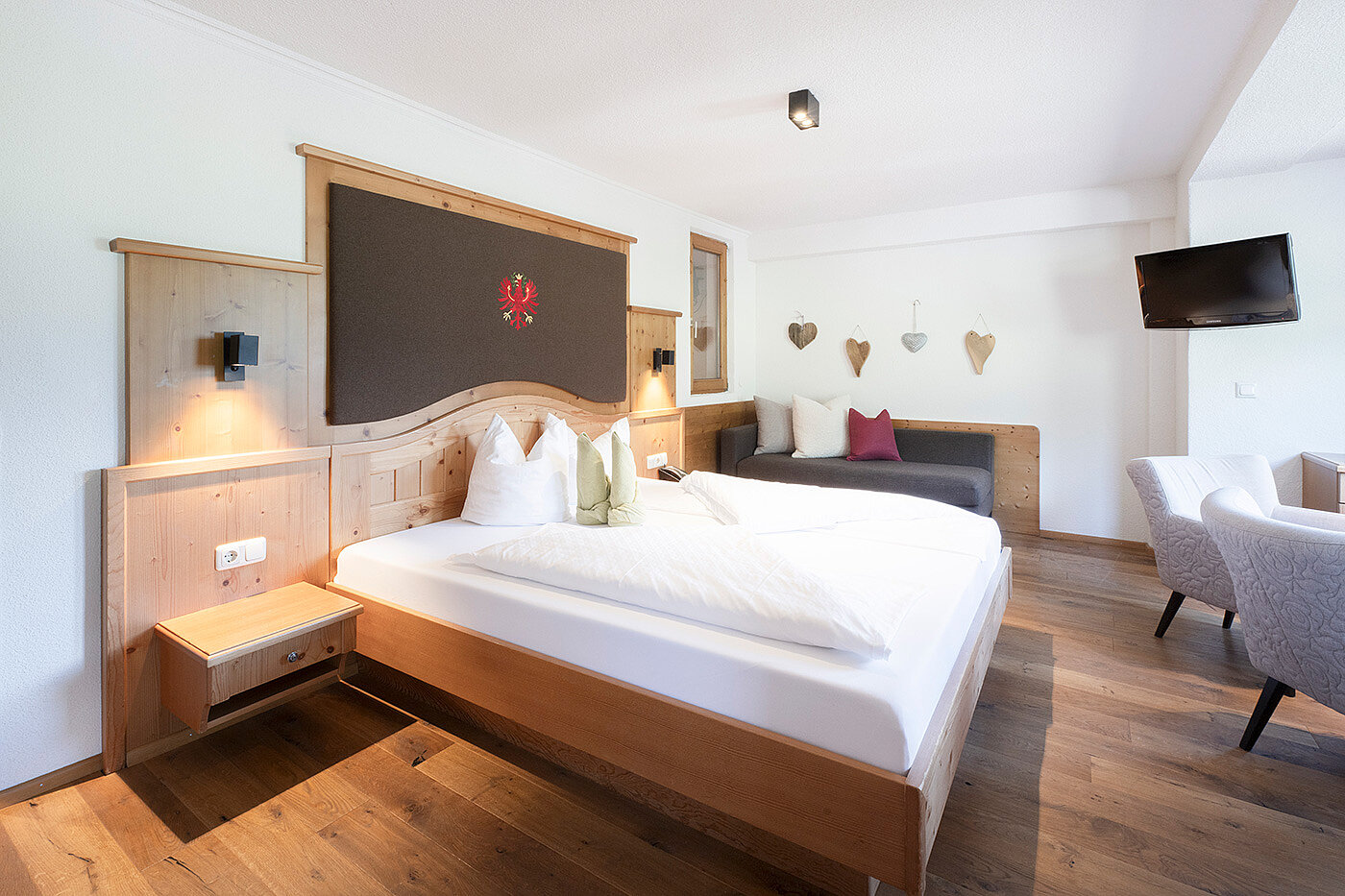 Helles Hotelzimmer mit großem Doppelbett, Holzmöbeln, Sofa und Wandfernseher an einer weißen Wand.