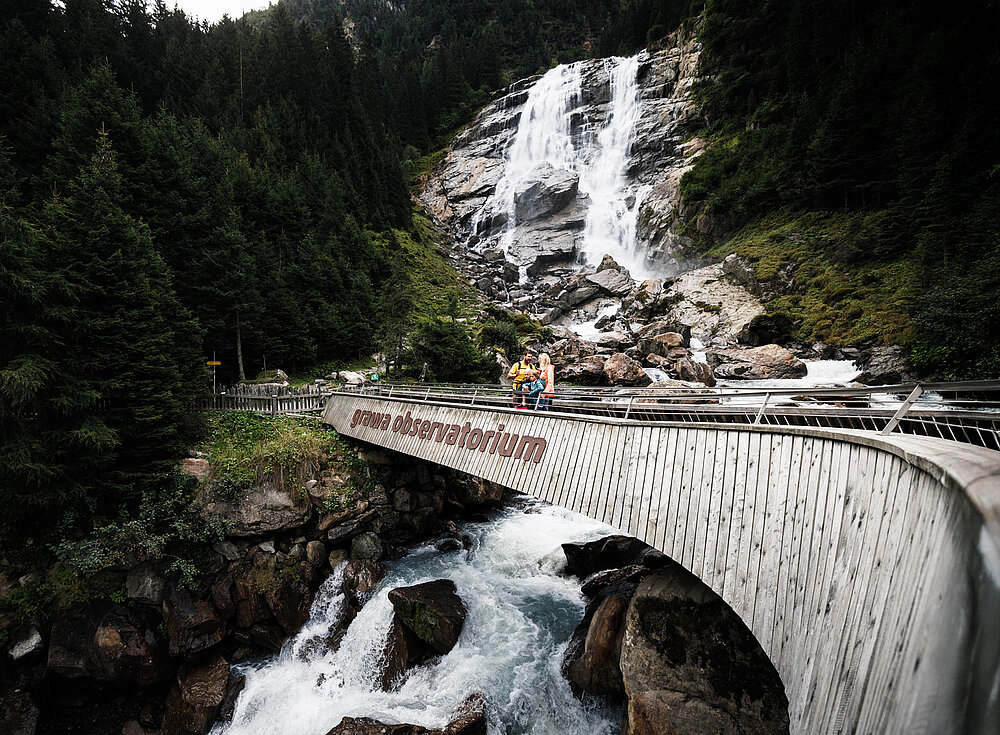 Eine Holzbrücke überquert einen felsigen Bach unterhalb eines hohen Wasserfalls, umgeben von dichten grünen Wäldern und Bergen.