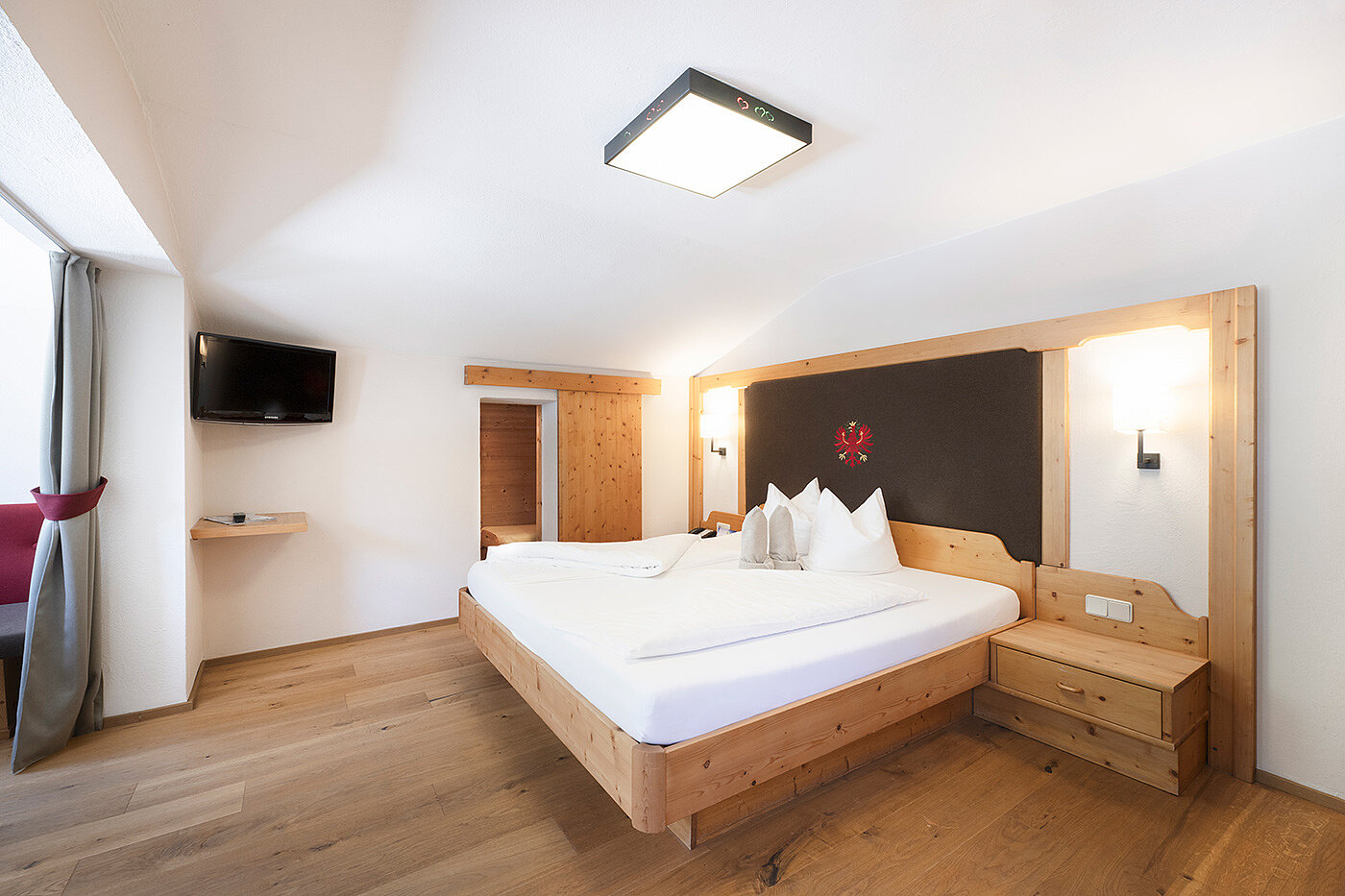 Helles Hotelzimmer mit großem Bett, Holzmöbeln, Fernseher an der Wand und einem Fenster mit Vorhängen.
