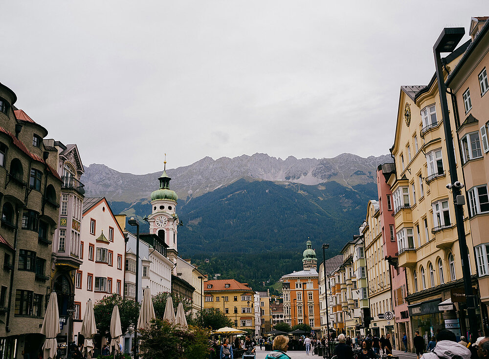 Eine Straße gesäumt von farbenfrohen Gebäuden und Cafés, mit Bergen und bewölktem Himmel im Hintergrund.