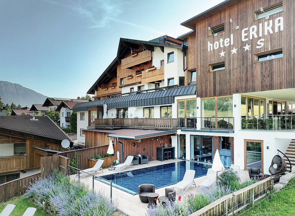 Außenansicht des Hotels Erika mit Holzfassade, Balkonen, Außenpool, Liegestühlen und Bergen im Hintergrund.