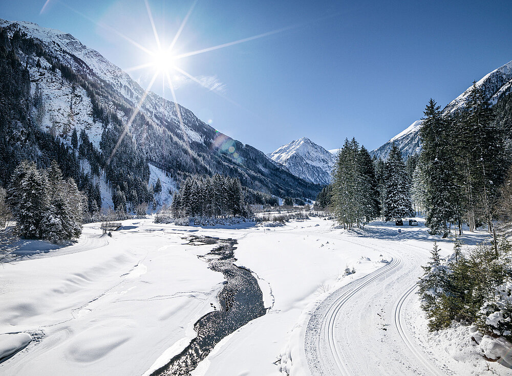 Schneebedecktes Tal mit einem Fluss, Kiefern, Bergen und strahlender Sonne an einem klaren blauen Himmel.