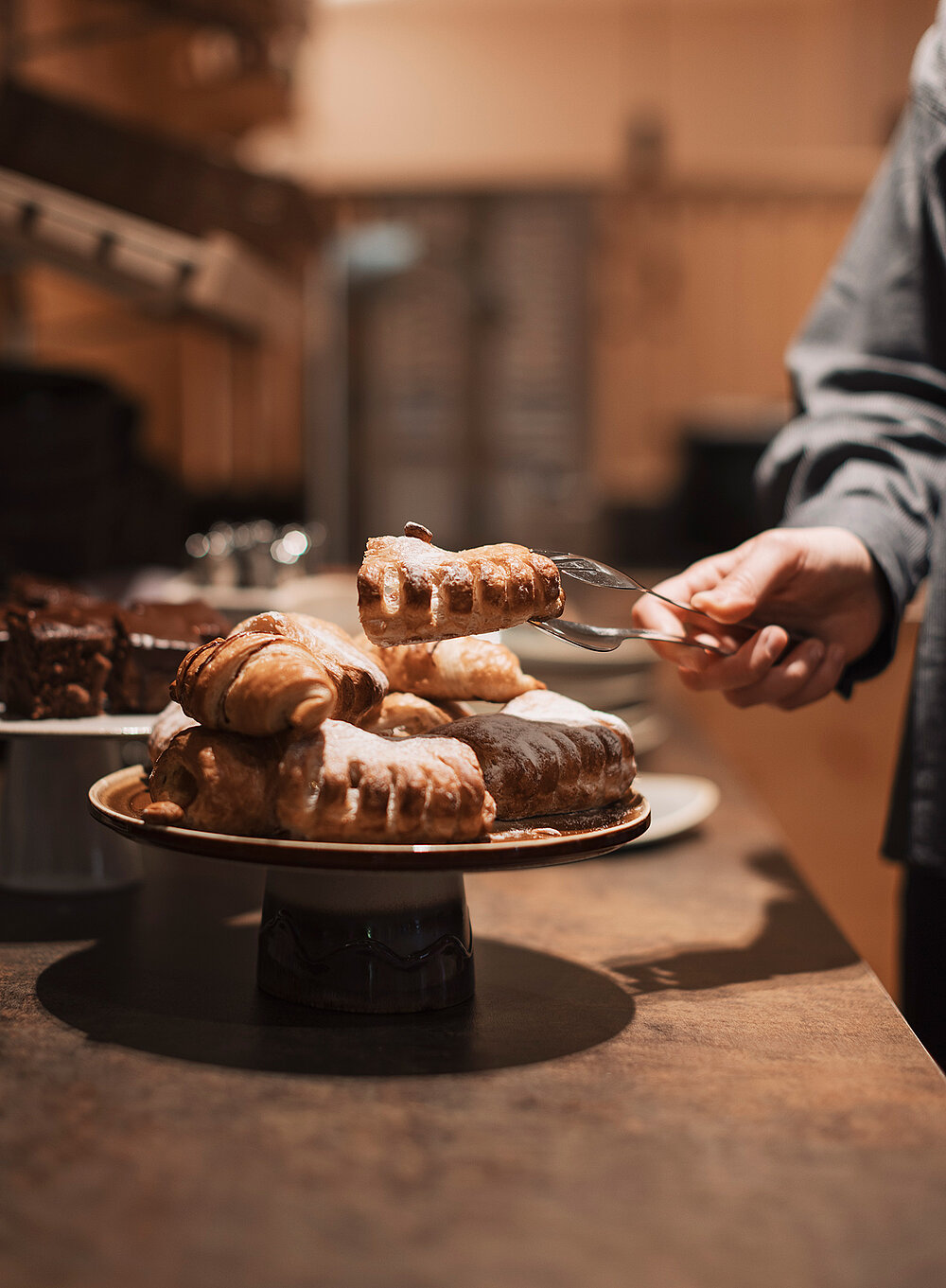 Eine Person serviert in einer warm beleuchteten Küche oder Bäckerei mit einer Zange Gebäck auf einem Teller.