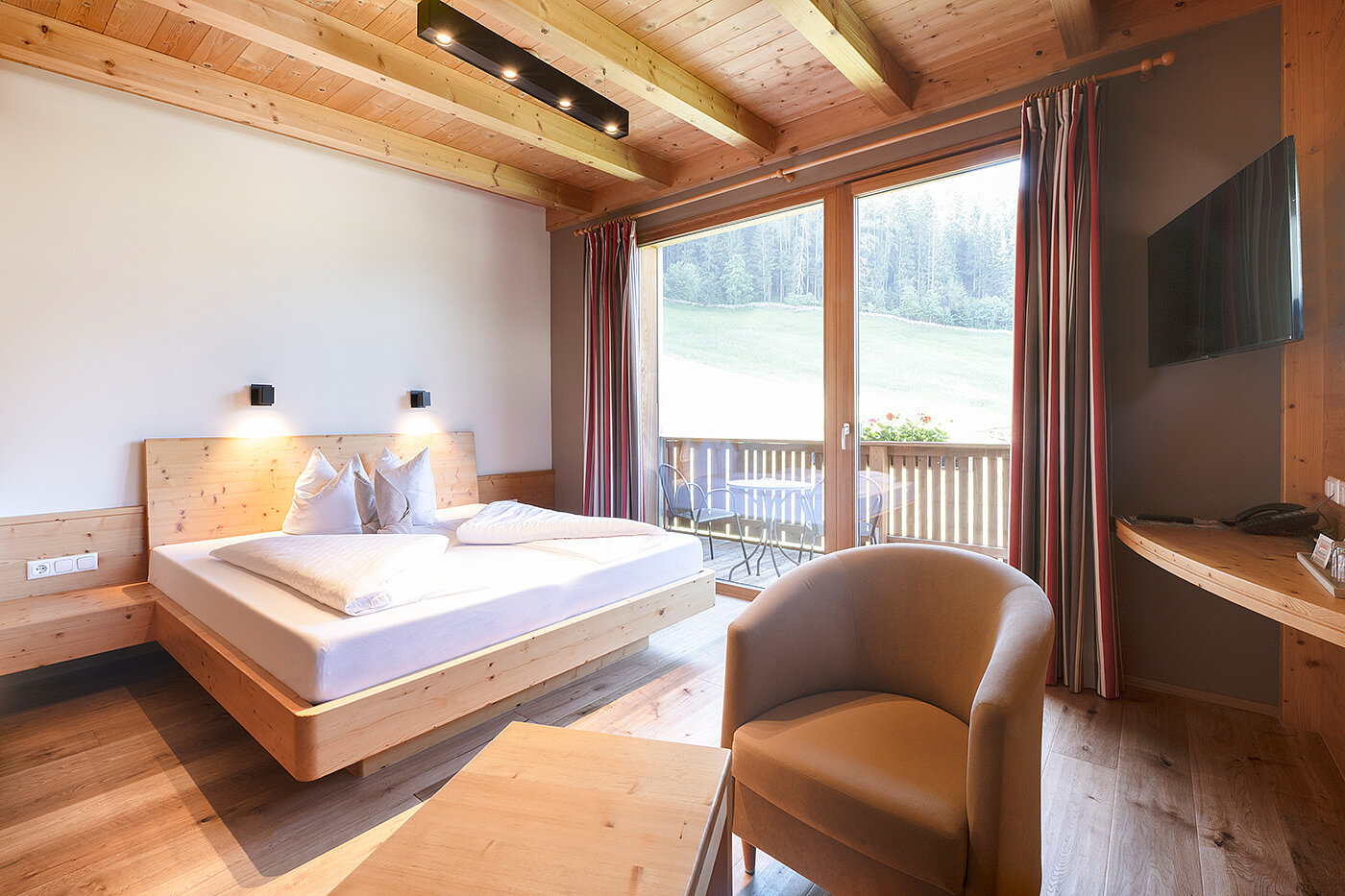 Modernes Schlafzimmer aus Holz mit großem Bett, Sessel, Fernseher und Balkon mit Blick auf eine Wiese und Bäume.