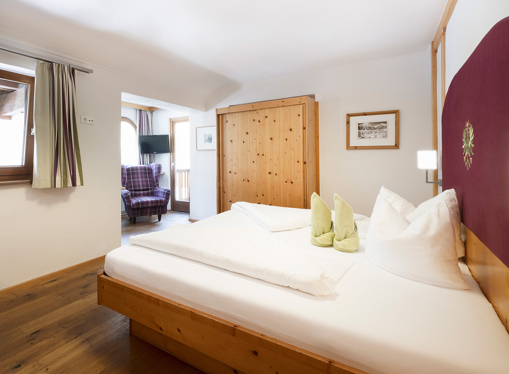 Helles Hotelzimmer mit großem Bett, Holzmöbeln, weißer Bettwäsche und einem Fenster mit hellen Vorhängen.