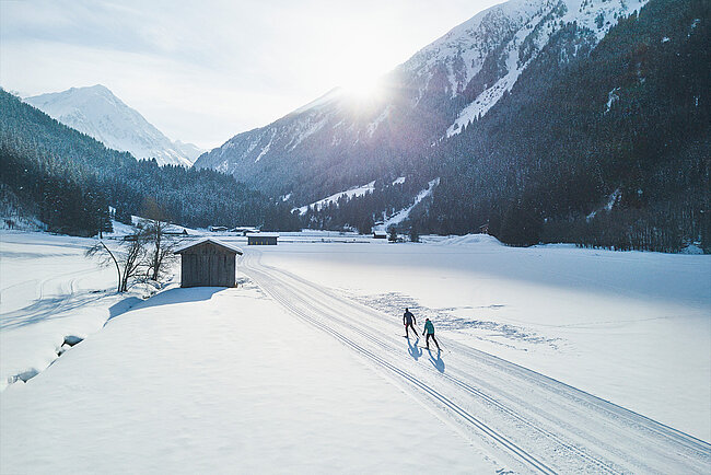 Zwei Personen fahren unter der strahlenden Wintersonne auf einer schneebedeckten Piste in der Nähe einer Hütte in einem verschneiten Bergtal Ski.